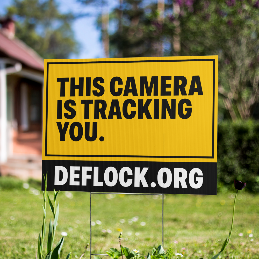 A yellow and black yard sign on a metal stake in a grassy yard, reading 'THIS CAMERA IS TRACKING YOU.' with 'DEFLOCK.