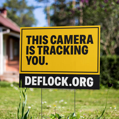 A yellow and black yard sign on a metal stake in a grassy yard, reading 'THIS CAMERA IS TRACKING YOU.' with 'DEFLOCK.